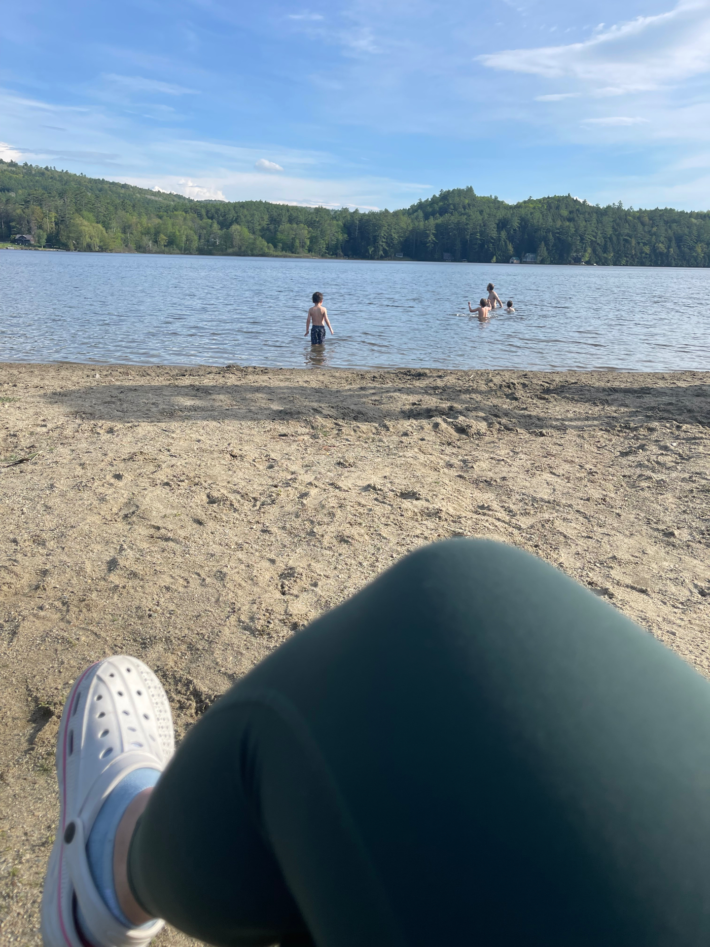 View from the sandy beach of kids wading in a calm Vermont lake with forested hills across the water