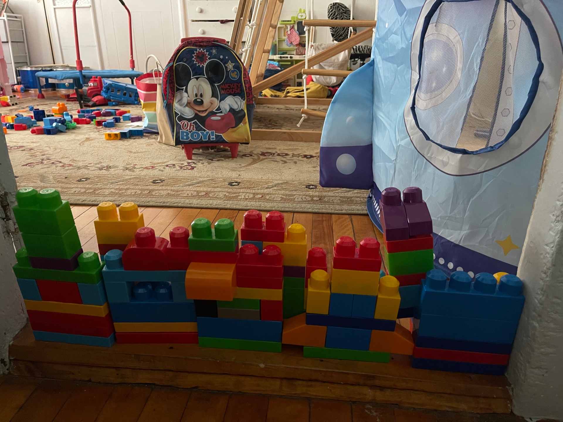 A long wall built from colorful mega blocks across the playroom floor with a Mickey Mouse backpack and rocket tent in the background